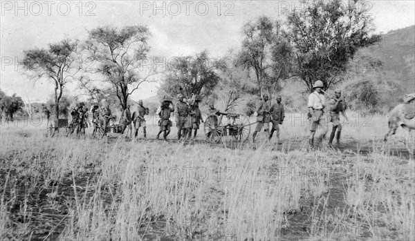 Machine-gun section of the King's African Rifles on trek. A European officer leads a line of African askaris (soldiers) as they pull a small gun carriage through scrubby landscape. Mozambique, 1918. Mozambique, Southern Africa, Africa.