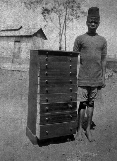 African cabinetmaker. An employee of the Public Works Department, identified in the the caption as 'Makongoro Wa Kusaga', stands beside a tallboy or chest of narrow drawers he has made. He wears a government uniform that displays the initials 'PWD' on the chest. British East Africa (Kenya), January 1916. Kenya, Eastern Africa, Africa.