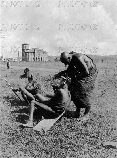 Kikuyu hairdresser. A Kikuyu man dresses the hair of another man seated on a plank on the ground. In the background can be seen the General Post Office of Nairobi. Nairobi, British East Africa (Kenya), 1911. Nairobi, Nairobi Area, Kenya, Eastern Africa, Africa.