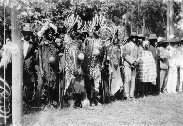 African leaders at the North Kavirondo show. African leaders await presentation to the Governor of Kenya at the North Kavirondo agricultural show. They wear headdresses decorated with split hippopotamus tusks, animal hides and hold ostrich eggs. European onlookers can be seen in the background. Mumias, Kenya, January 1931. Mumias, West (Kenya), Kenya, Eastern Africa, Africa.