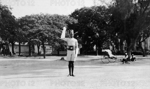Kenyan traffic policeman. The policeman signals 'stop' with his white glove. Behind him two rickshaw pullers squat beneath a tree, an empty rickshaw beside them. Kenya, circa 1928. Kenya, Eastern Africa, Africa.