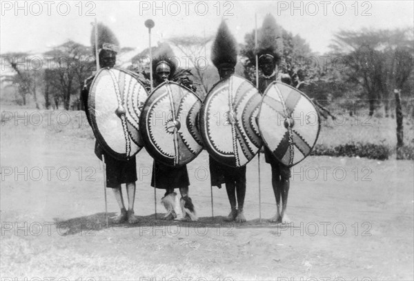 Nandi film extras. Four Nandi warriors recruited as film extras for the 1930 film 'Africa Speaks'. They wear elaborate traditional costumes and pose with shields, spears and staffs. Kapsabet, Kenya, 1928. Kapsabet, Rift Valley, Kenya, Eastern Africa, Africa.