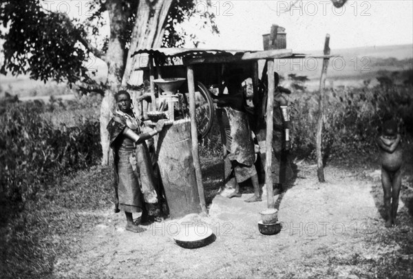 Nandi women grinding maize. Two Nandi women operate a small, partially-mechanized maize mill, which appears to involve a bicycle wheel. Near Kapsabet, Kenya, circa 1928. Kapsabet, Rift Valley, Kenya, Eastern Africa, Africa.