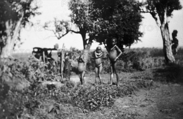 Nandi children. Several young, naked Nandi children stand in front of a small maize mill operated by adult women. Near Kapsabet, Kenya, circa 1928. Kapsabet, Rift Valley, Kenya, Eastern Africa, Africa.