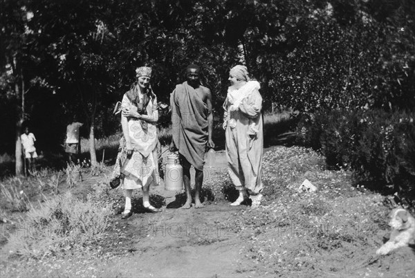 Christmas fancy dress with servant. Two European women wearing fancy dress stand either side of an African domestic servant carrying a milk can along a garden path. The woman on the right is the wife of Charles Bungey. Machakos, Kenya, December 1926. Machakos, East (Kenya), Kenya, Eastern Africa, Africa.