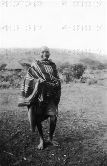 Kamba boy. Informal portrait of a smiling Kamba boy wearing a striped blanket. Kenya, circa 1927. Machakos, East (Kenya), Kenya, Eastern Africa, Africa.