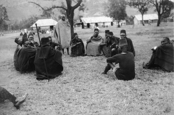 Policeman meets with Kamba elders. A Kenyan policeman sits on the ground for a meeting with a group of elders ('wazee') in traditional dress. Machakos, Kenya, 1927. Machakos, East (Kenya), Kenya, Eastern Africa, Africa.