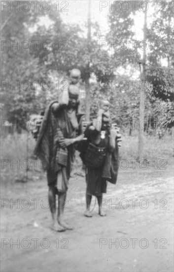 Kamba women with children. Two barefooted Kamba women in traditional dress carry small children on their shoulders along a dirt road. Machakos, Kenya, June 1927. Machakos, East (Kenya), Kenya, Eastern Africa, Africa.