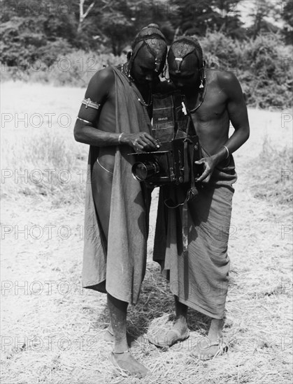Looking through the viewfinder. Two African men, possibly Maasai, peer into the viewfinder of a large format single lens reflex Graflex camera that they hold between them. They are dressed in traditional costume and jewellery and their braided hair is tied back and daubed with ochre. Amboseli, Tanganyika Territory (Tanzania), 1 October 1951. Amboseli, Kilimanjaro, Tanzania, Eastern Africa, Africa.
