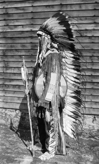 Native American costume. A European man dressed in traditional native American costume including a long feathered headdress and tassled clothing. He carries a shield under his arm and holds a stick decorated with feathers. Canada, circa 1922., Alberta, Canada, North America, North America .