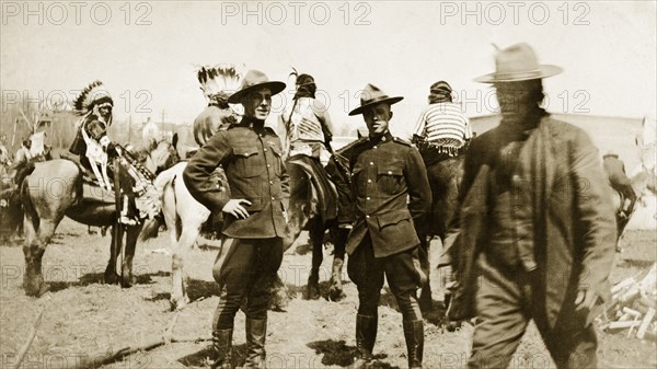 Royal Canadian Mounted Police. Two uniformed Royal Canadian Mounted Policemen at a native American camp. Behind them, several native Amercian men wearing traditional dress wait mounted on their horses. Canada, circa 1925. Canada, North America, North America .