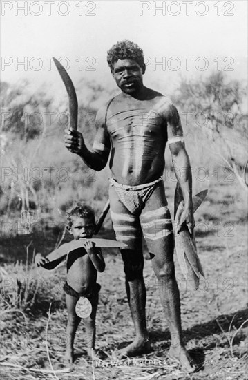 Aborigines with boomerangs. An aboriginal man holding several wooden boomerangs poses for the camera with a small child. The man has stripes painted onto his body and displays numerous horizontal scar lines on his chest. North West Australia, 1925., West Australia, Australia, Australia, Oceania.