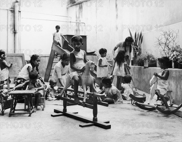 Sri Lankan nursery. Three women supervise a group of children at play in a well-equipped Methodist nursery. The two children in the foreground ride wooden rocking horses whilst others can be seen playing with a variety of toys including a blackboard and a slide. Ceylon (Sri Lanka), 1957. Sri Lanka, Southern Asia, Asia.