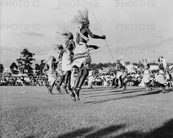 Watusi dancers at the Kampala Trade Show. Male Watusi dancers wearing traditional costume and elaborate feathered headdresses, are captured mid-jump as they perform a dance for a crowd of African spectators at the Kampala trade show. Kampala, Kenya, 15-18 December 1955. Kampala, Central (Uganda), Uganda, Eastern Africa, Africa.