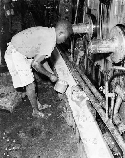 Testing juice at a sugar mill. An African worker dips a testing pot into a trough of juice being processed at the Miwani Sugar Mills. Miwani, Kenya, 7-8 November 1955. Miwani, Nyanza, Kenya, Eastern Africa, Africa.