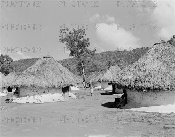 Huts for sugar mill labourers. People seek shade under the thatched roofs of a number of round huts at a camp assigned to workers employed by the Miwani Sugar Mills. Miwani, Kenya, 7-8 November 1955. Miwani, Nyanza, Kenya, Eastern Africa, Africa.