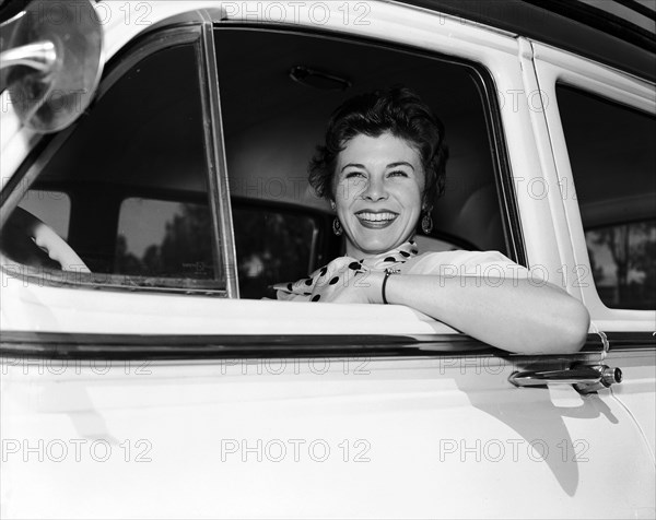 Daphne Dale in the driving seat. Dancer Daphne Dale smiles from the driver's seat of a car. She wears a spotted scarf and rests her elbow out of the open window. Kenya, 11 November 1955. Kenya, Eastern Africa, Africa.