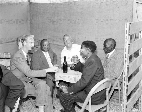 Tusker beer stand at the Royal Show. African and European visitors drink from glasses around a table at the Tusker stand at the annual Kampala trade show. Kampala, Uganda, 15-18 December 1955. Kampala, Central (Uganda), Uganda, Eastern Africa, Africa.