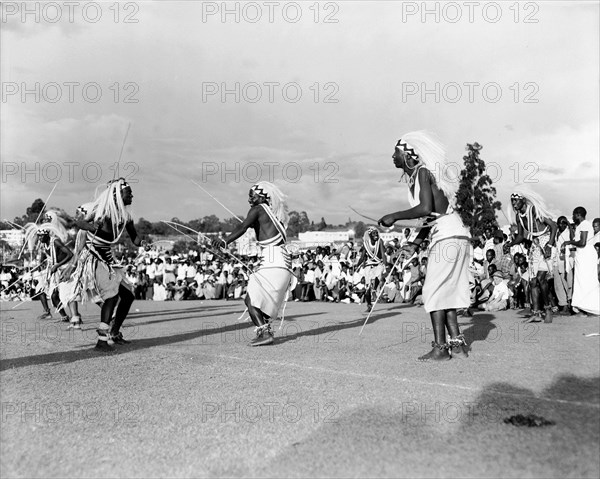 Watusi dancers at the Kampala Trade Show. Male Watusi dancers wearing traditional costume and elaborate feathered headdresses, perform a dance with sticks for a crowd of African spectators at the Kampala trade show. Kampala, Uganda, 15-18 December 1955. Kampala, Central (Uganda), Uganda, Eastern Africa, Africa.