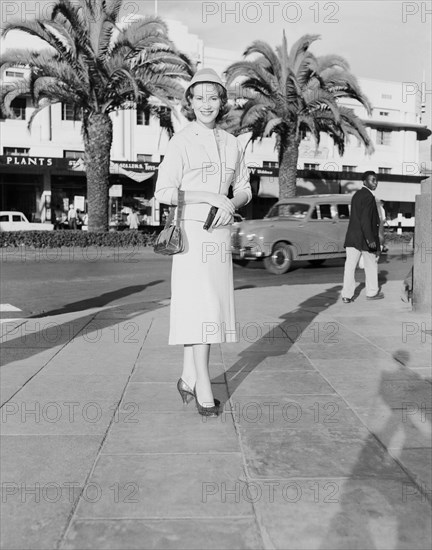 Fashion 'shoot'. Publicity shot for Regent outfitters. Fashion model Ino poses for the camera in the centre of town wearing a Regent outfit. She carries a handbag in one hand and casually holds a revolver in the other. Kenya, 14 October 1955. Kenya, Eastern Africa, Africa.
