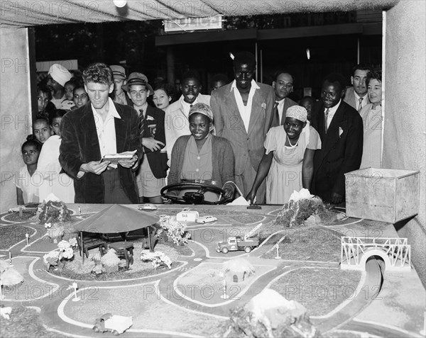 Driving game. A mixed race crowd gathers around a detailed miniature road model at the Motor Sports Club stand at the Royal Show. An African woman holds a steering wheel and appears to be controlling the movement of the model cars. Nairobi, Kenya, 28 September-1 October 1955. Nairobi, Nairobi Area, Kenya, Eastern Africa, Africa.