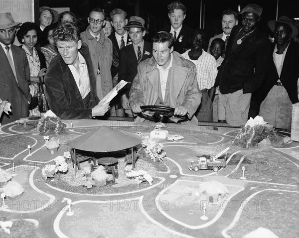Driving game. A mixed race crowd gathers around a detailed miniature road model at the Motor Sports Club stand at the Royal Show. An European man holds a steering wheel and appears to be controlling the movement of the model cars. Nairobi, Kenya, 28 September-1 October 1955. Nairobi, Nairobi Area, Kenya, Eastern Africa, Africa.