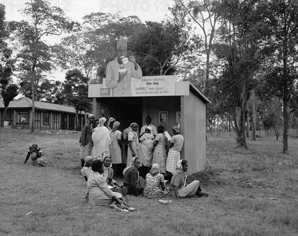 Knorr-Swiss stall at the Royal Show. A group of African women congregate around a Knorr-Swiss stall at the Royal Show. A cut-out sign above them shows the cartoon of a smiling man drinking Knorr-Swiss clear soup. Nairobi, Kenya, 28 September-1 October 1955. Nairobi, Nairobi Area, Kenya, Eastern Africa, Africa.