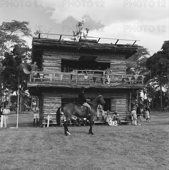 Reporters box at the SJAK show. A rider in the class 14 gambler stakes pauses in front of a reporter's box decorated with bunting and megaphones at the SJAK show (Sports Journalists Association of Kenya). Kenya, 20 August 1955. Kenya, Eastern Africa, Africa.