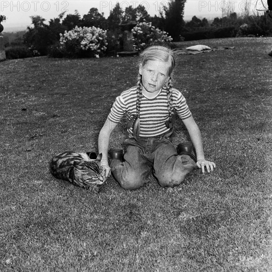 In the Russel's garden. A young girl dressed in jeans with her hair braided into two long plaits, plays with a tabby cat in the Russel's garden. Kenya, 4 September 1955. Kenya, Eastern Africa, Africa.