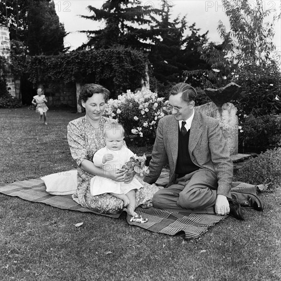 In the Russel's garden. A couple sitting on a rug in the Russel family's garden entertain a baby with flowers and beads as a young girl runs across the lawn in the background. Kenya, 4 September 1955. Kenya, Eastern Africa, Africa.