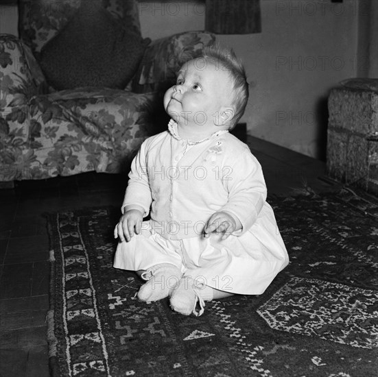 Baby at the Russel's house. A baby sitting on a patterned rug at the Russel's family house looks expectantly upwards. Kenya, 4 September 1955. Kenya, Eastern Africa, Africa.