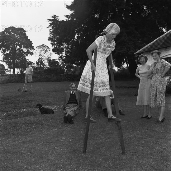 Miss Russel walks on stilts. The Russel's daughter attempts to walk on wooden stilts in the family garden, much to the amusement of onlooking family and friends. Kenya, 4 September 1955. Kenya, Eastern Africa, Africa.