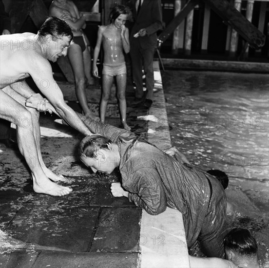 Swimming gala antics. A man in a suit is helped out of a swimming pool by three others wearing swimwear at a swimming gala social. Kenya, 17 September 1955. Kenya, Eastern Africa, Africa.