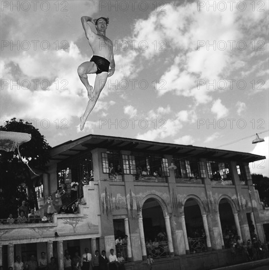 Crazy diving. A man wearing trunks at a swimming gala launches himself into the air from a diving board, striking a pose as he descends. A crowd of onlookers watch the spectacle from a large colonial-style building. Kenya, 17 September 1955. Kenya, Eastern Africa, Africa.