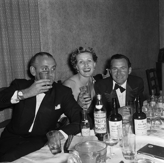 Partying at the SJAK dance. A group dressed in formal evening wear raise their glasses at a dance held for the Sports Journalists Association of Kenya (SJAK). Bottles of Haig's gold label whisky and Hennessy's cognac can be seen in the foreground and the table is littered with empty glasses. Kenya, 19 August 1955. Kenya, Eastern Africa, Africa.