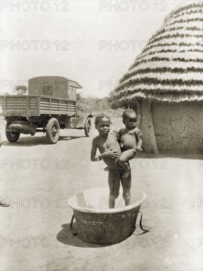 Child and baby in bath tub. A semi-naked child stands inside an empty bath tub, holding a baby on one hip outside a village hut with a thatched roof. An open-backed truck is parked in the background. Sudan, circa 1930. Sudan, Eastern Africa, Africa.