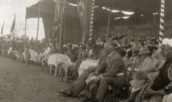 Kings' Day celebrations. A large, mostly male, crowd of onlookers sit around tables beneath a decorative cloth canopy at an event celebrating Kings' Day. Africans in turbans sit next to Europeans in solatopis and the union jack and flag of Islam are displayed in unison. Wadi Halfa, Sudan, 1932. Wadi Halfa, North (Sudan), Sudan, Eastern Africa, Africa.