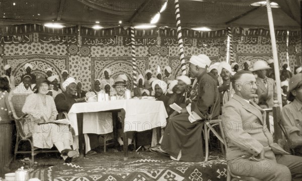 Kings' Day celebrations. A large, mostly male, crowd sit around tables beneath an elaborately decorative cloth canopy at an event celebrating Kings' Day. Africans wearing turbans and robes share tables with Europeans wearing suits and solatopis. Wadi Halfa, Sudan, 1932. Wadi Halfa, North (Sudan), Sudan, Eastern Africa, Africa.