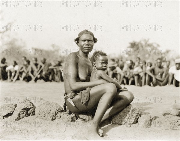 Sleeping sickness inspection. A Sudanese woman and her baby sit in front of a crowd of Sudanese people lined up awaiting inspection for sleeping sickness by a senior medical officer. Sudan, February 1929. Sudan, Eastern Africa, Africa.