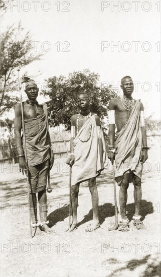 Three Nuer men. Three young Nuer men wear traditional dress and carry sticks. They are adorned with jewellery including armlets, anklets, beaded necklaces and decorative head bands. Sudan, circa 1930. Sudan, Eastern Africa, Africa.