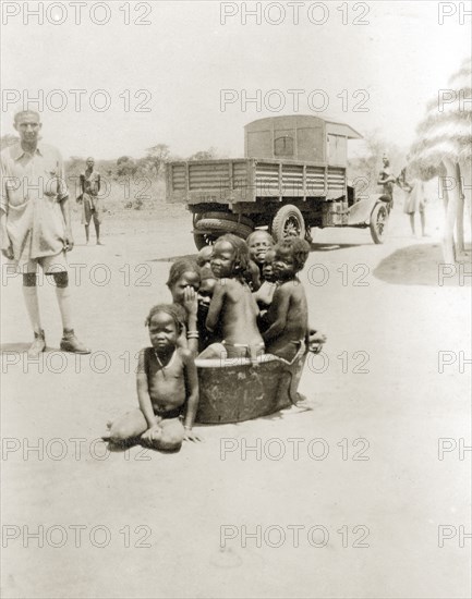 Children in bath tub. A group of semi-naked African children squash together in an empty bath tub outside a village hut with a thatched roof. An open-backed truck is parked in the background. Sudan, circa 1930. Sudan, Eastern Africa, Africa.