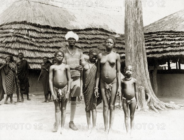 Saleh and Balanda girls. An African man wearing a turban with shorts and long socks stands outside a thatched village hut with a group of semi-naked Balanda girls. Sudan, circa 1930. Sudan, Eastern Africa, Africa.