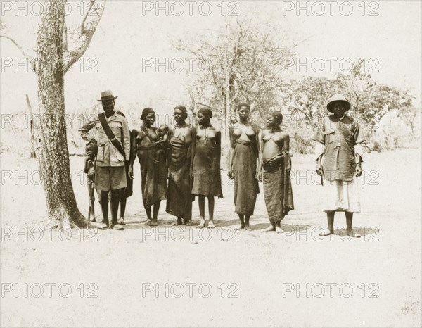 Balanda chief and wives. A Balanda chief and his deputy with their six wives. Both men wear European-style hats and sashes, pinned with badges that read 'Chief'. The women wear traditional dress with short braided hair and are naked from the waist up. Sudan, circa 1925. Sudan, Eastern Africa, Africa.