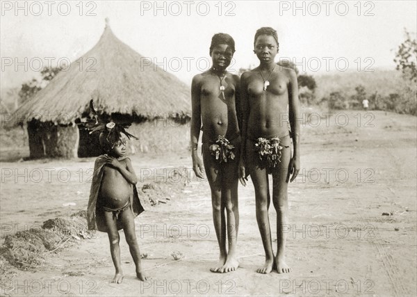 Balanda children on the Bo road. Three semi-naked Balanda children on the road to Bo town. The older girls wear necklaces and bunches of leaves that cover their genitalia: the young boy wears a cape and an elaborate headdress made from feathers. Sudan, circa 1930., West Bahr el Ghazal, Sudan, Eastern Africa, Africa.