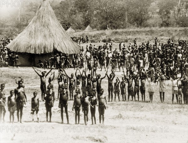 Sleeping sickness patients. A crowd of sleeping sickness patients wave at the camera from a camp containing thatched huts. Yubo, Sudan, circa 1930. Yubo, West Bahr el Ghazal, Sudan, Eastern Africa, Africa.