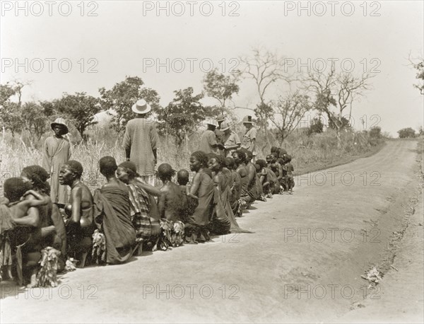 Sleeping sickness inspection. Sudanese women and babies sit in a line on a roadside awaiting inspection for sleeping sickness by a senior medical officer. Sudan, February 1929. Sudan, Eastern Africa, Africa.