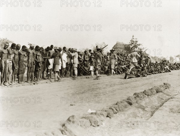 Sleeping sickness inspection. Sudanese men and boys stand and sit in a line on a roadside awaiting inspection for sleeping sickness by a senior medical officer. Armed African and European colonial officials in uniforms oversee the crowd. Sudan, February 1929. Sudan, Eastern Africa, Africa.