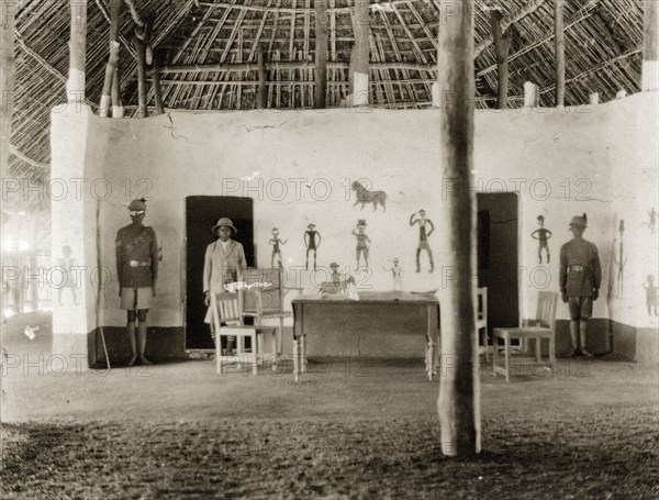 The DC's new office. African colonial officials in uniform stand beside a desk and chairs inside a large thatched hut identified as 'the DC's new office'. Crudely drawn figures can be seen painted onto the plaster wall behind them. Tambura, Sudan, circa 1930., West Equatoria, Sudan, Eastern Africa, Africa.