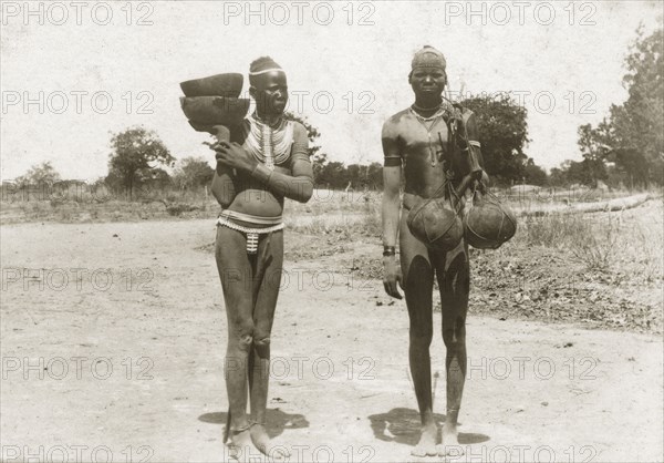 Dinkas on a country road. A young Dinka man and woman carry bowls and gourds on a country road. Naked apart from their jewellery, they both wear head bands: the woman displaying an ornate beaded necklace and shell belt. Sudan, circa 1930. Sudan, Eastern Africa, Africa.