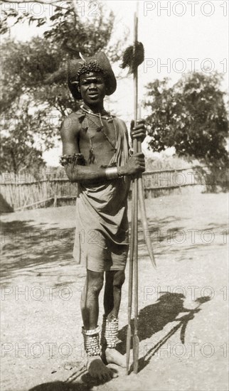 A Shilluk man. A Shilluk man pictured outdoors wears traditonal dress comprising beaded jewellery decorated with feathers, armlets and anklets. He holds two spears and displays a striking hat and headband. Sudan, circa 1930., Upper Nile, Sudan, Eastern Africa, Africa.